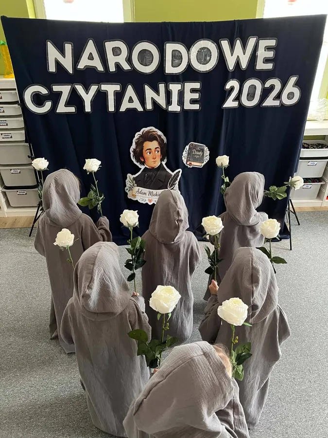 Dzieci w szarych pelerynach z białymi różami przed dekoracją Narodowe Czytanie 2026