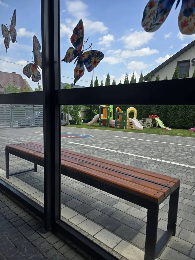 Ławka pod wiatą z motylami na szybie, w tle plac zabaw przedszkola