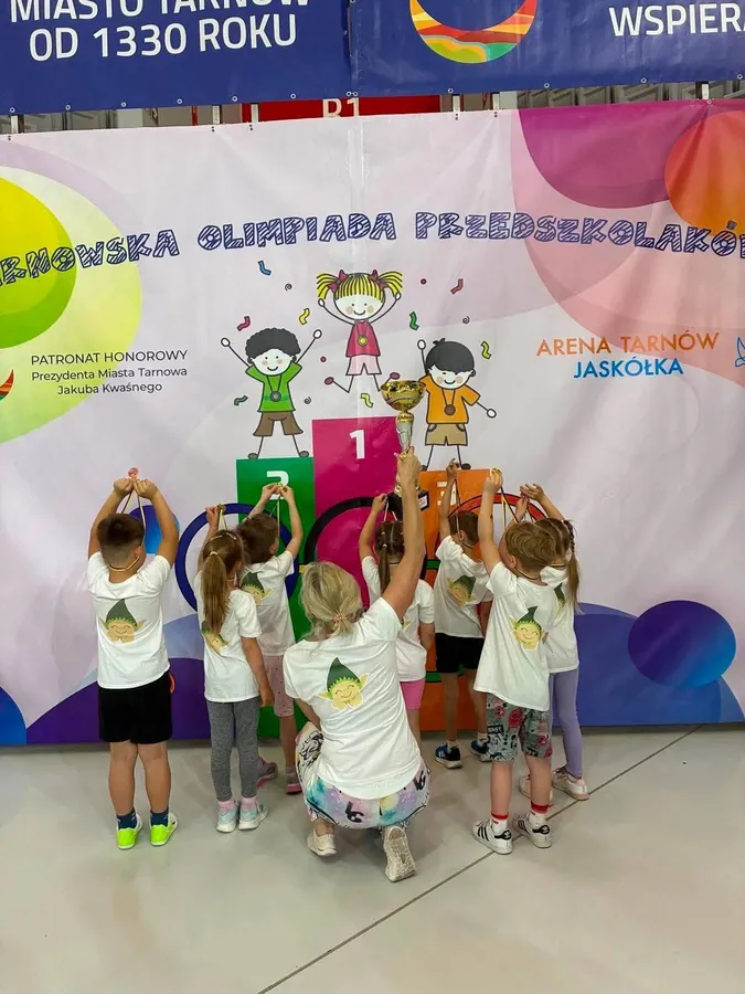 Przedszkolaki w białych koszulkach przed ścianką Tarnowskiej Olimpiady Przedszkolaków